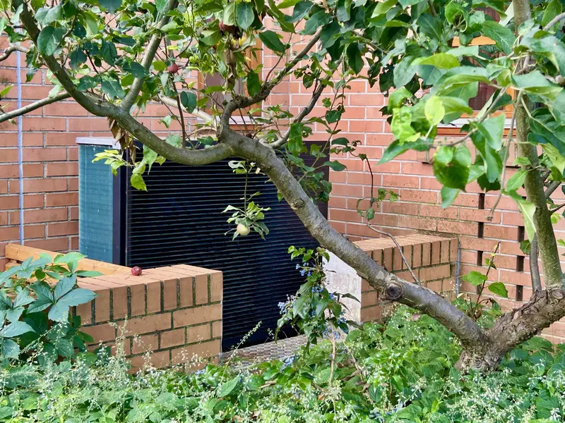 Wärmepumpe vor roter Backsteinwand, hinter Apfelbaum