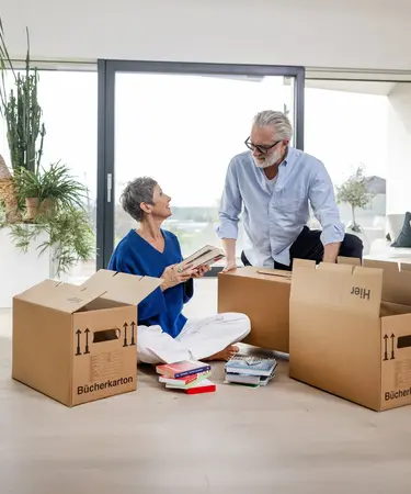 Älteres Paar sitzt am Boden im Wohnzimmer und packt Umzugskartons