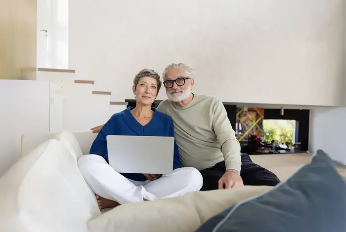 Bestager Pärchen auf Couch mit Laptop. Blick in die Kamera