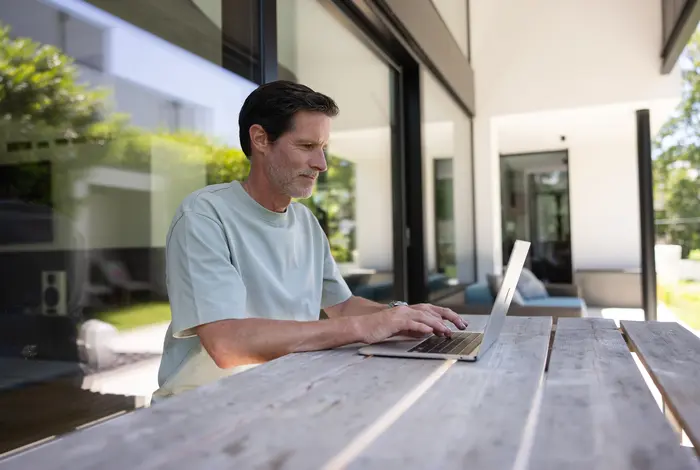 Mann sitz an einem Holzterrassentisch und arbeitet am Laptop.