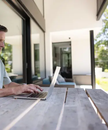 Mann sitz an einem Holzterrassentisch und arbeitet am Laptop.