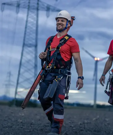 Zwei Netz NÖ Mitarbeiter gehen über ein Feld, im Hintergrund ein Hochspannungsleitung und ein Windrad in der Abenddämmerung.