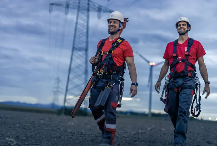 Zwei Netz NÖ Mitarbeiter gehen über ein Feld, im Hintergrund ein Hochspannungsleitung und ein Windrad in der Abenddämmerung.