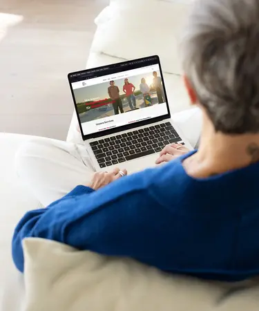 Ältere Frau sitzt mit Laptop am Sofa. Blick über die Schulter auf ihren Screen.