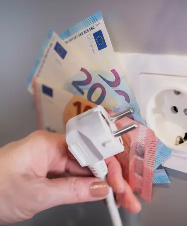 Hand hält Stromstecker und aufgefächerte Euroscheine vor einer Steckdose.