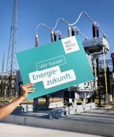 Eine Männerhand hält ein türkises "Energiezukunft"-Schuld in die Höhe. Im Hintergrund das modernisierte Umspannwerk Haag Süd.