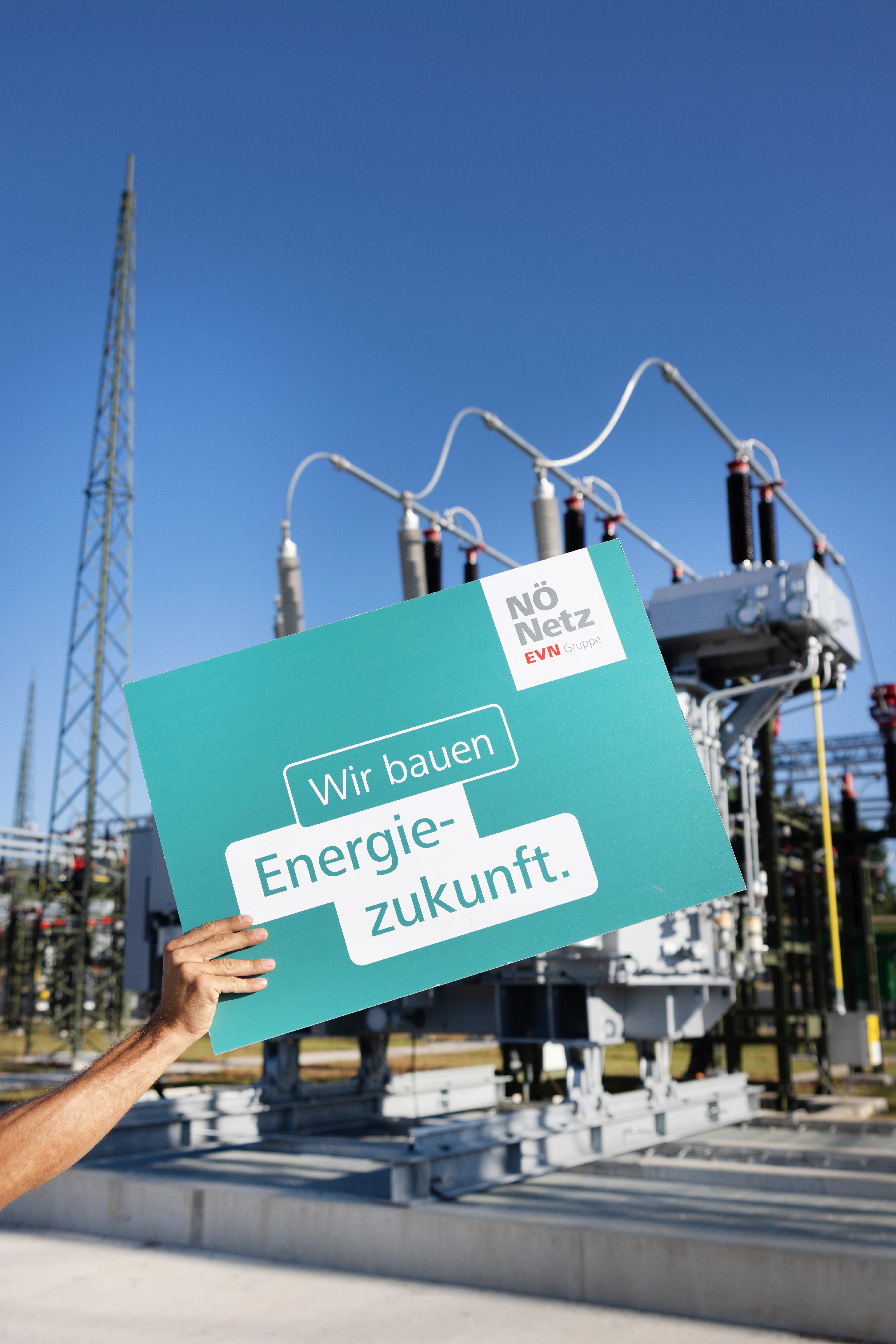 Eine Männerhand hält ein türkises "Energiezukunft"-Schuld in die Höhe. Im Hintergrund das modernisierte Umspannwerk Haag Süd.