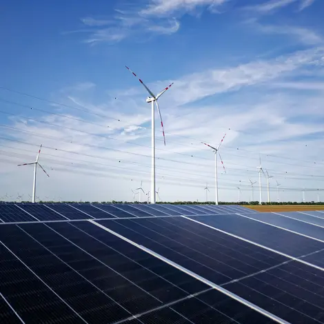 Im Vordergrund liegt eine Flächen PV-Anlage vor Windrädern. Im Hintergrund eine Stromleitung.