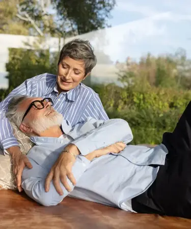 Älteres Paar in Wintergarten: Frau sitzt über ihn gebeugt, Mann liegt mit Kopf an sie gelehnt. 