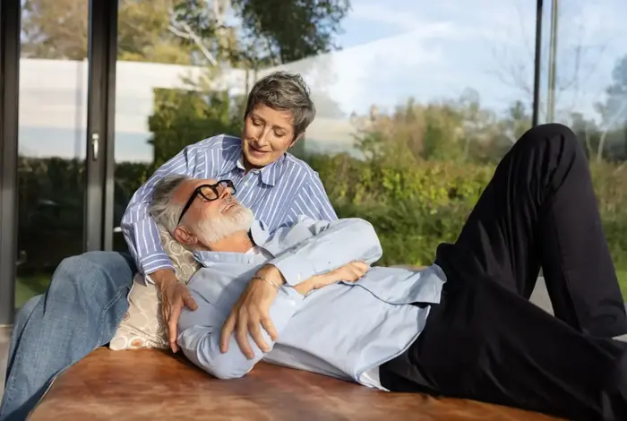 Älteres Paar in Wintergarten: Frau sitzt über ihn gebeugt, Mann liegt mit Kopf an sie gelehnt. 